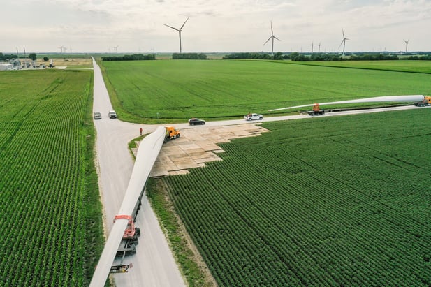 Wind turbine blade delivery across a stabilized access road constructed with matting solutions, enabling safe transport through agricultural terrain for renewable energy infrastructure projects.