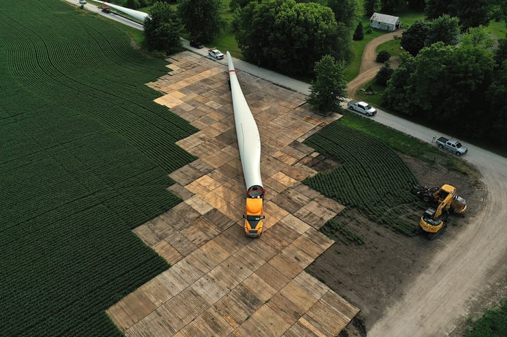 Wind turbine blade delivery across a stabilized access road constructed with matting solutions, enabling safe transport through agricultural terrain for renewable energy infrastructure projects.